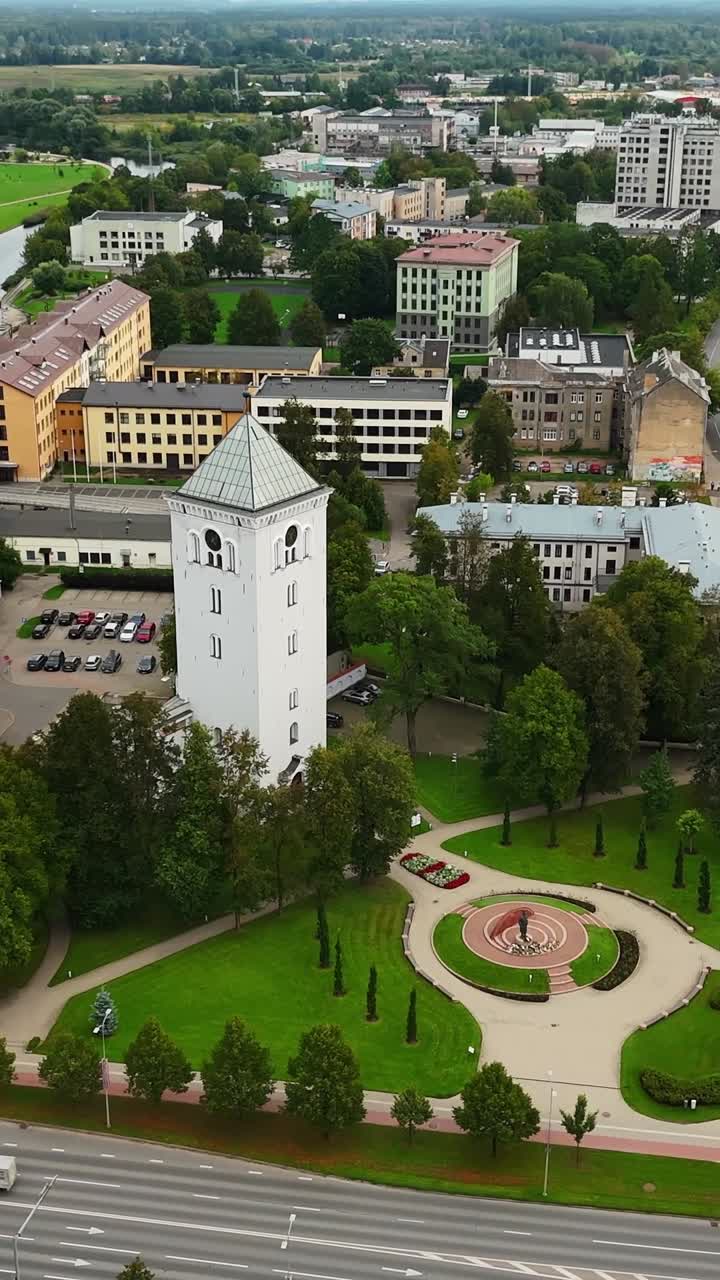 Vertical drone view of Jelgava, Latvia—Holy Trinity Church Tower anchors the scene, with Orthodox Cathedral in background, framed by roads, green spaces, and mixed architecture