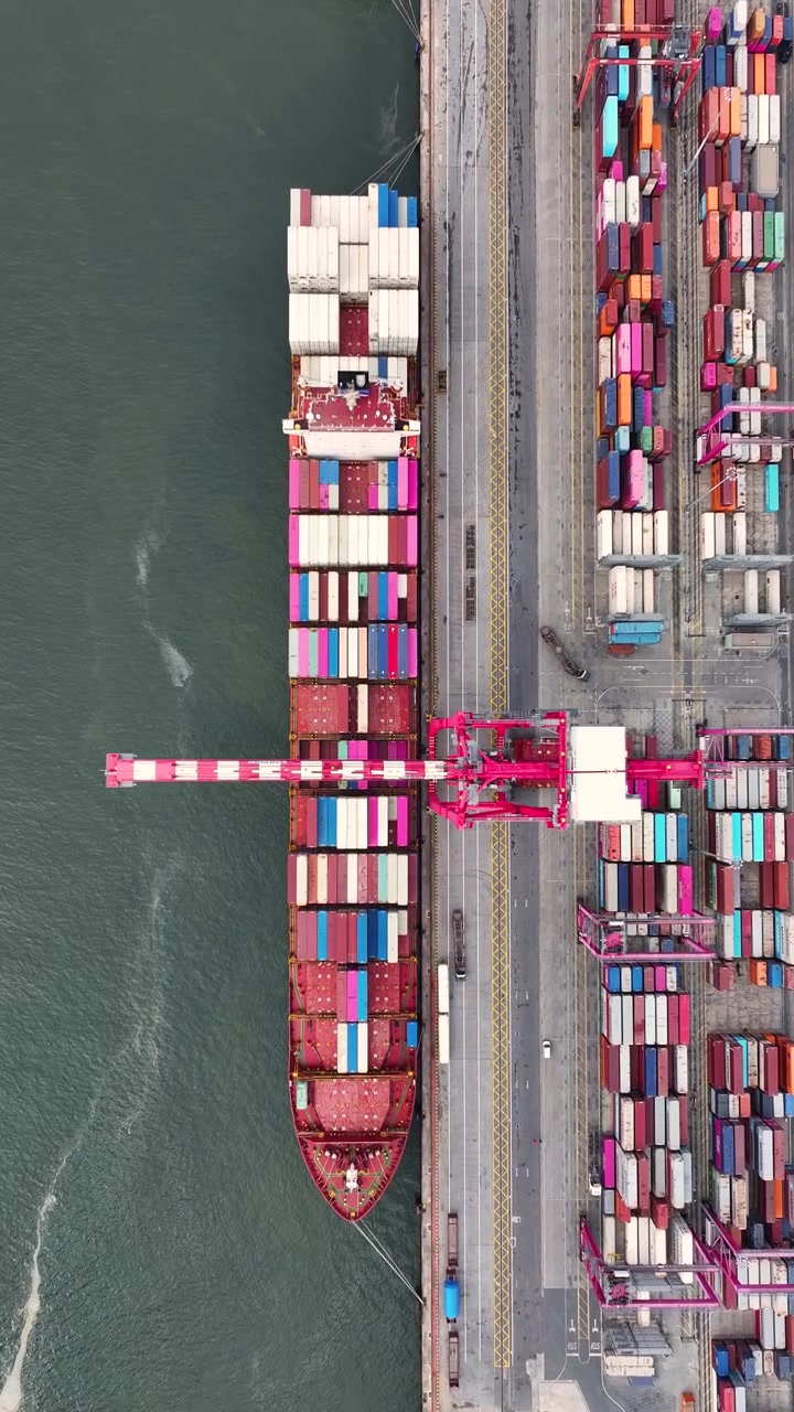 A busy container terminal with numerous shipping containers stacked on the dock. A large container ship is being loaded with cargo while trucks transport goods nearby.