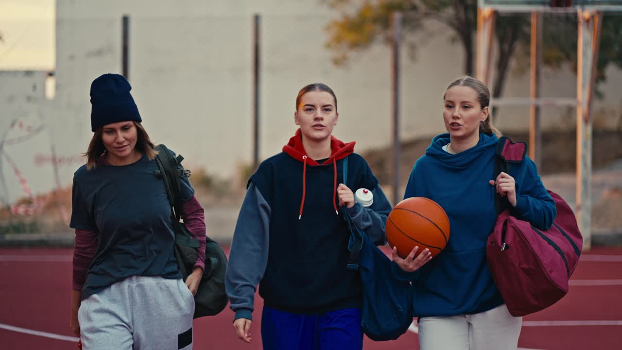 Three confident blonde girls in sportswear with an orange basketball walk along a red street court in the summer morning. Happy blonde girls go to basketball training and game in summer