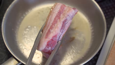 Top-down-side-view-of-pork-belly-being-cooked-inside-stainless-steel-pan,-moved-around-with-tongs