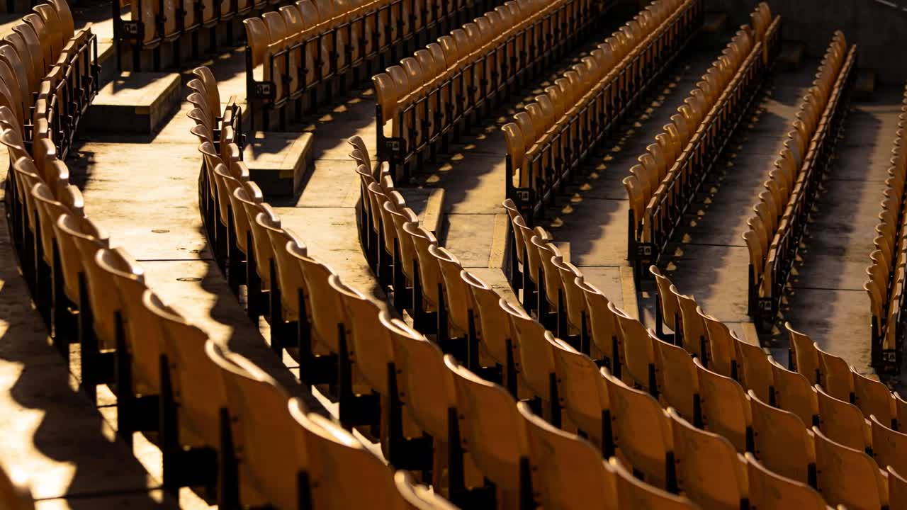 Camera panning stadium fold-down seats on concrete tiers, revealing supports, stair treads, shadows