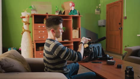 man arranges camera gear on table in home studio, adjusting lens and microphone. cozy workspace with green walls, sofa, and storage shelves. focused on video production setup