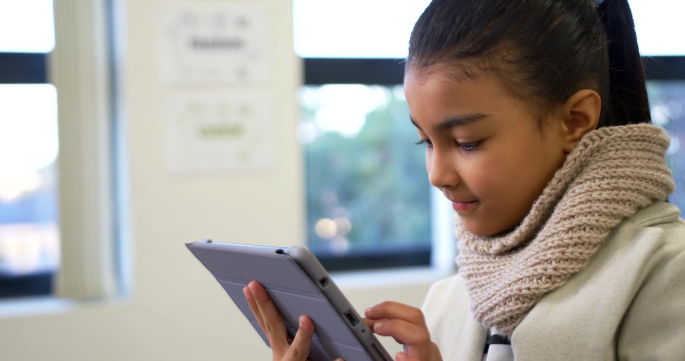 Pre-teen African American girl holding tablet and tapping screen by classroom window, wearing scarf