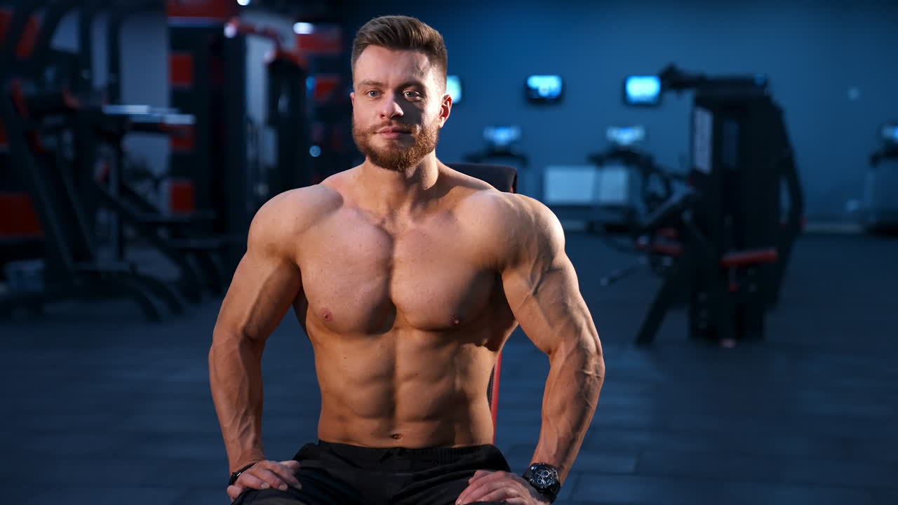 Male sportsman with naked torso and big muscles sits on training chair in modern gym. Male beauty concept, Slow motion.
