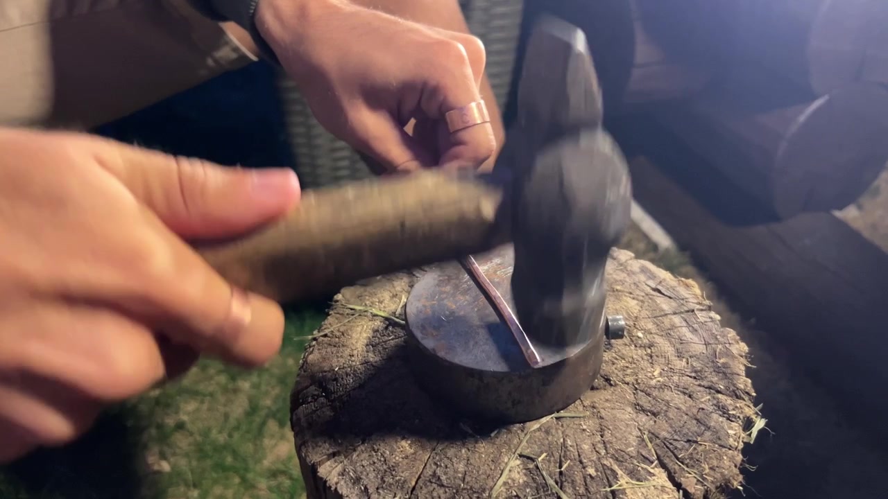 Close-up shot of hands using a hammer to forge copper wire on a small anvil outdoors at night. Ideal for artisan crafts, DIY tutorials, and metalworking videos.