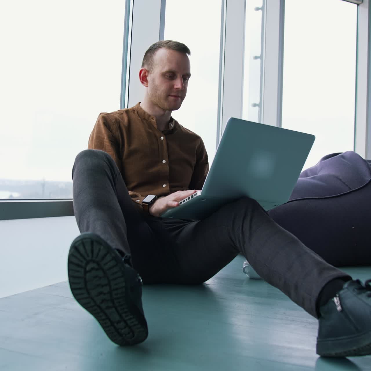 Young freelancer working on a laptop. Man in shirt and trousers sitting on a floor and using a gadget to work in office. Panoramic windows view