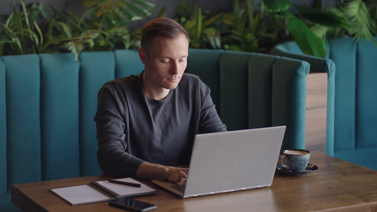 un joven con camisa está sentado en una mesa con una computadora portátil y escribiendo en el teclado. un estudiante puede estudiar a distancia. un hombre de negocios lleva a cabo su negocio a distancia