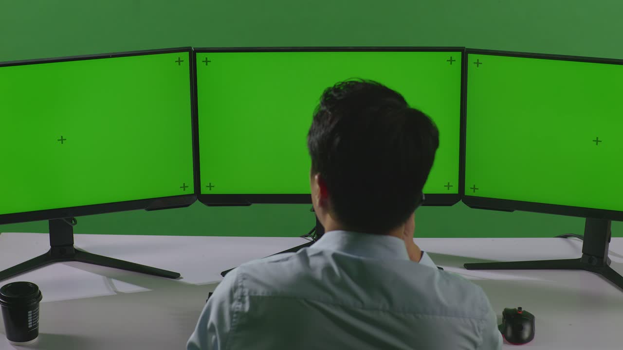녹색 스크린 스튜디오에서 모의 여러 컴퓨터 모니터와 함께 작업하는 동안 휴대 전화로 이야기하는 아시아 남자의 뒷면