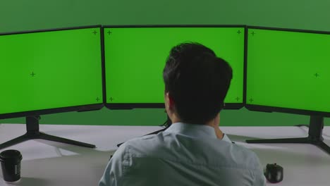 녹색 스크린 스튜디오에서 모의 여러 컴퓨터 모니터와 함께 작업하는 동안 휴대 전화로 이야기하는 아시아 남자의 뒷면