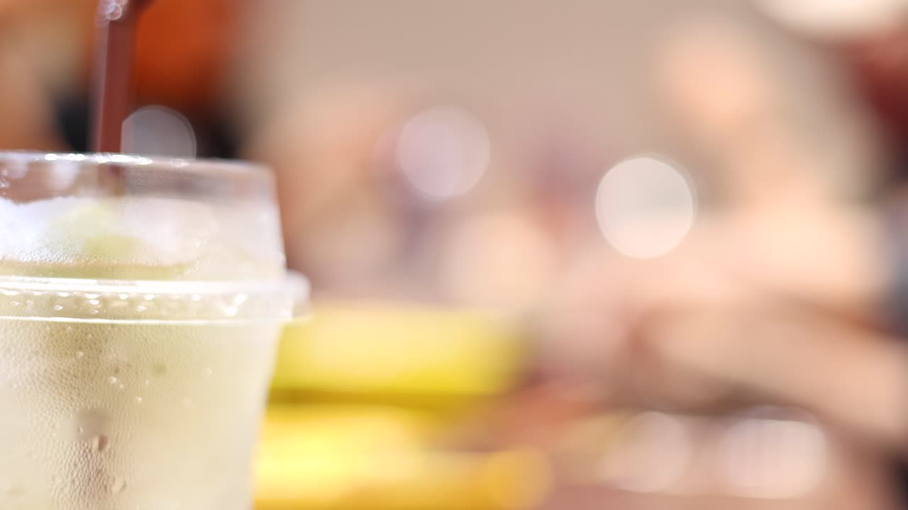 A close-up view of a soft drink being poured into a plastic cup filled with ice, with a blurred indoor restaurant background and warm lighting