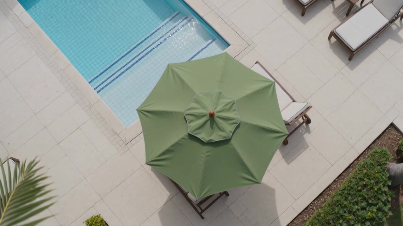 Telecamera aerea inclinata che rivela un ombrellone verde centrale sopra le sedie a bordo piscina per mostrare la disposizione.