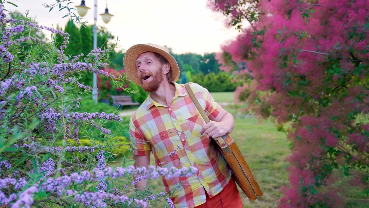 Artista felice con cappello di paglia che annusa fiori viola in un giardino colorato
