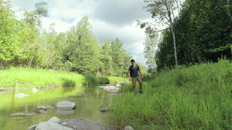 Einsamer-Moderner-Männlicher-Wanderer-In-Der-Wildnis,-Der-Inmitten-Einer-Waldlandschaft-Mit-Felsen-Und-üppig-Grünem-Sommerlaub-Ein-überwuchertes-Flussufer-Sucht