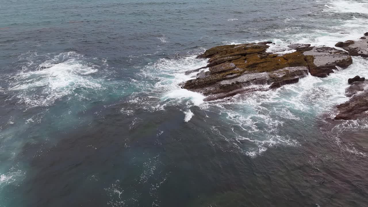 Waves swirl and crash gently against dark coastal rocks in a peaceful aerial ocean scene