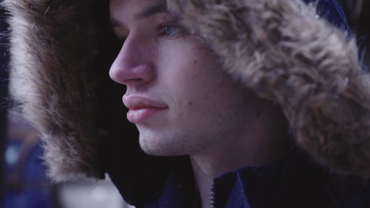vista lateral de un hombre joven y guapo con una chaqueta cálida con capucha peluda con nieve cayendo