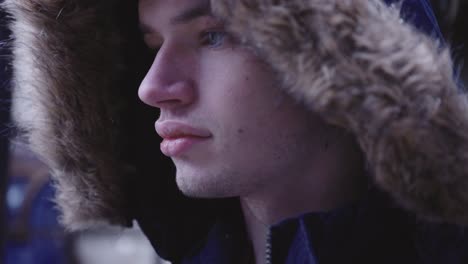 Side-View-Of-A-Young-And-Handsome-Man-Wearing-A-Warm-Jacket-With-Furry-Hood-With-Snow-Falling