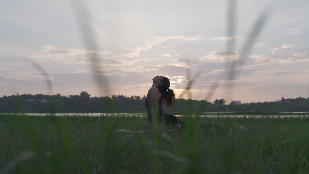 A wide-angle shot of a South Asian woman leaning back in a deep yoga pose on green grass, framed by soft sunrise light through tall blades