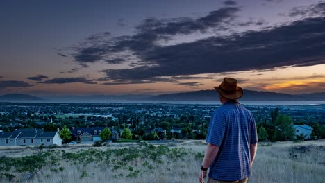 Ein-Mann-Steht-Ehrfürchtig-Da,-Während-Ein-Zeitraffer-Von-Tag-Zu-Nacht-Herauszoomt,-Um-Szenen-Der-Fernen-Berge-Und-Einer-Stadt-In-Einem-Tal-Mit-Einem-See-Im-Hintergrund-Zu-Enthüllen