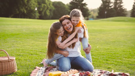 Two-Cute-Little-Sisters-Embracing-Her-Happy-Mother-From-Back-At-Park-During-Picnic