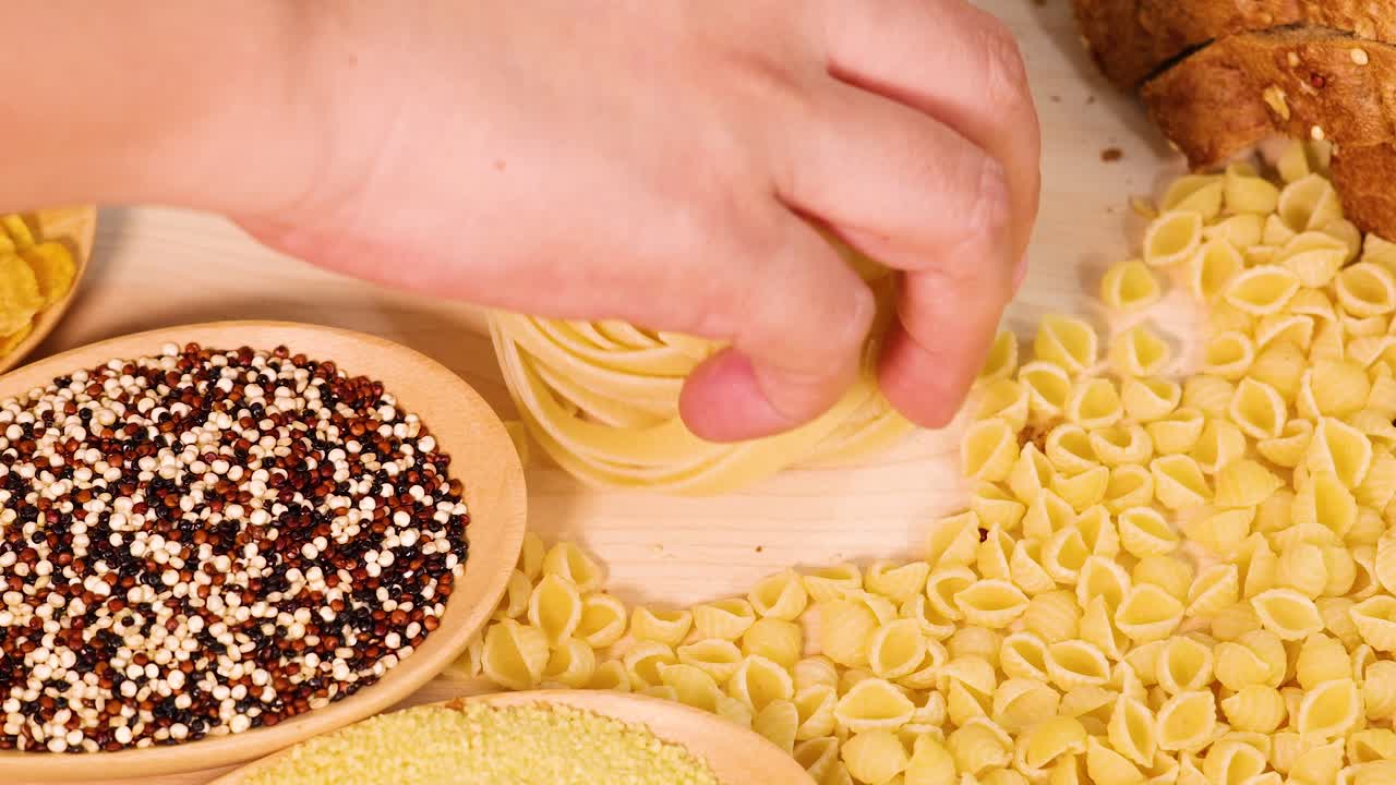 A hand arranges various pasta shapes and grains on a wooden surface under warm lighting, creating a visually appealing composition