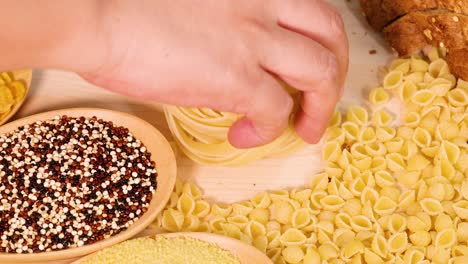 a hand arranges various pasta shapes and grains on a wooden surface under warm lighting, creating a visually appealing composition