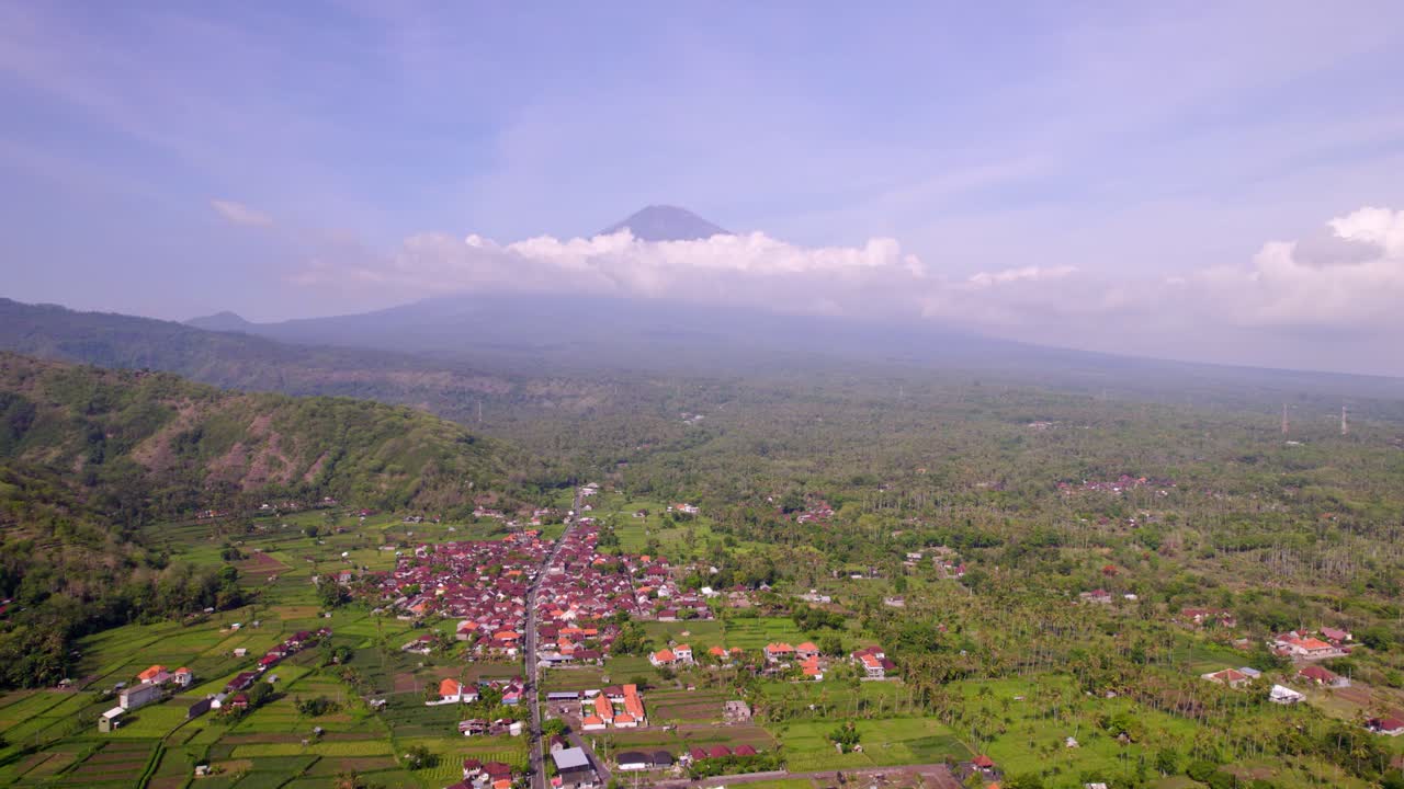 A paisagem rural de Amed, Bali, se estende abaixo com o Monte Agung e terreno verde em camadas, acompanhamento aéreo para a direita.