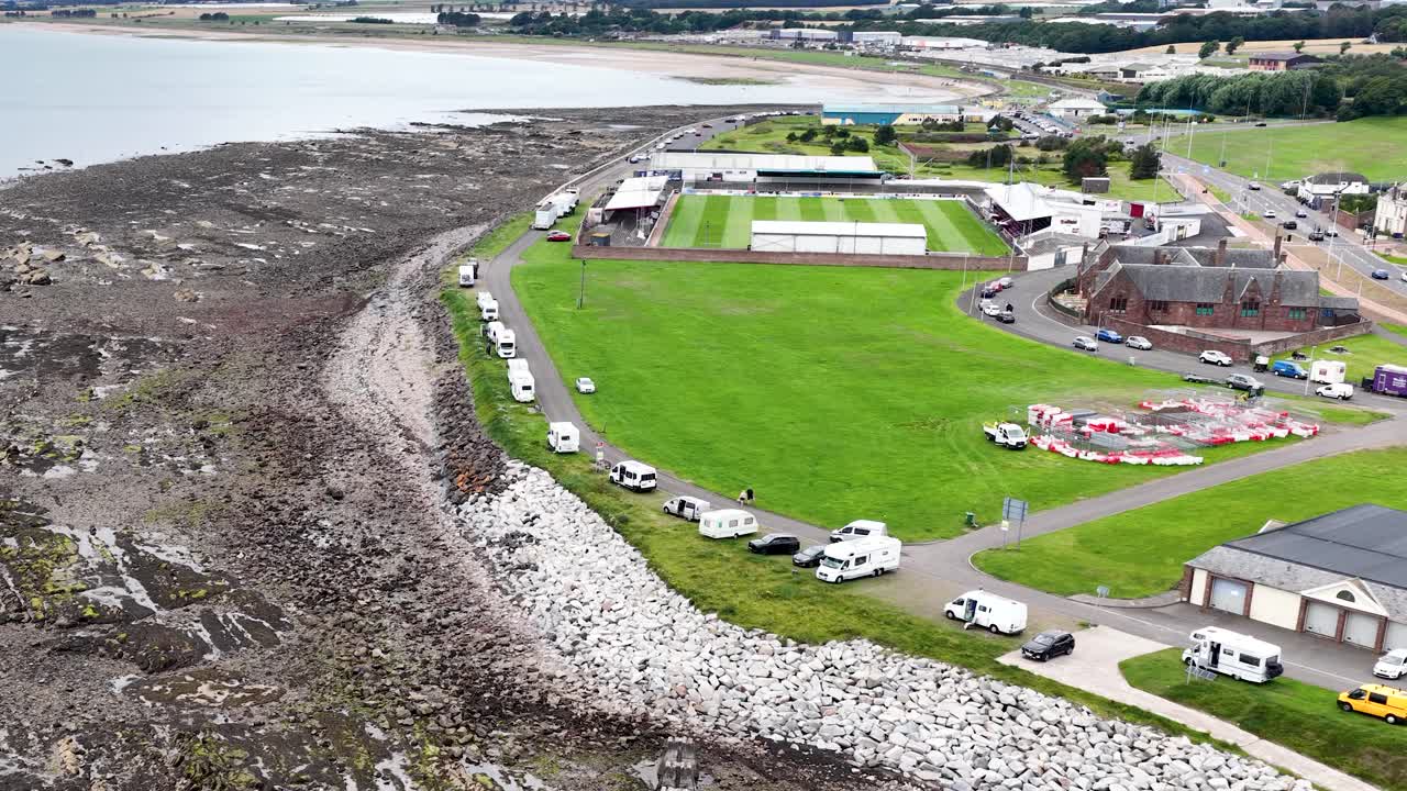 Drone glides above rocky shore, grassy fields, and buildings under bright daylight in Dundee, Scotland