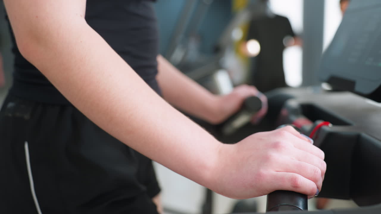 close up side view of woman in black athletic outfit holding treadmill handles during indoor workout session with blurred background of other gym users and fitness equipment under bright lighting
