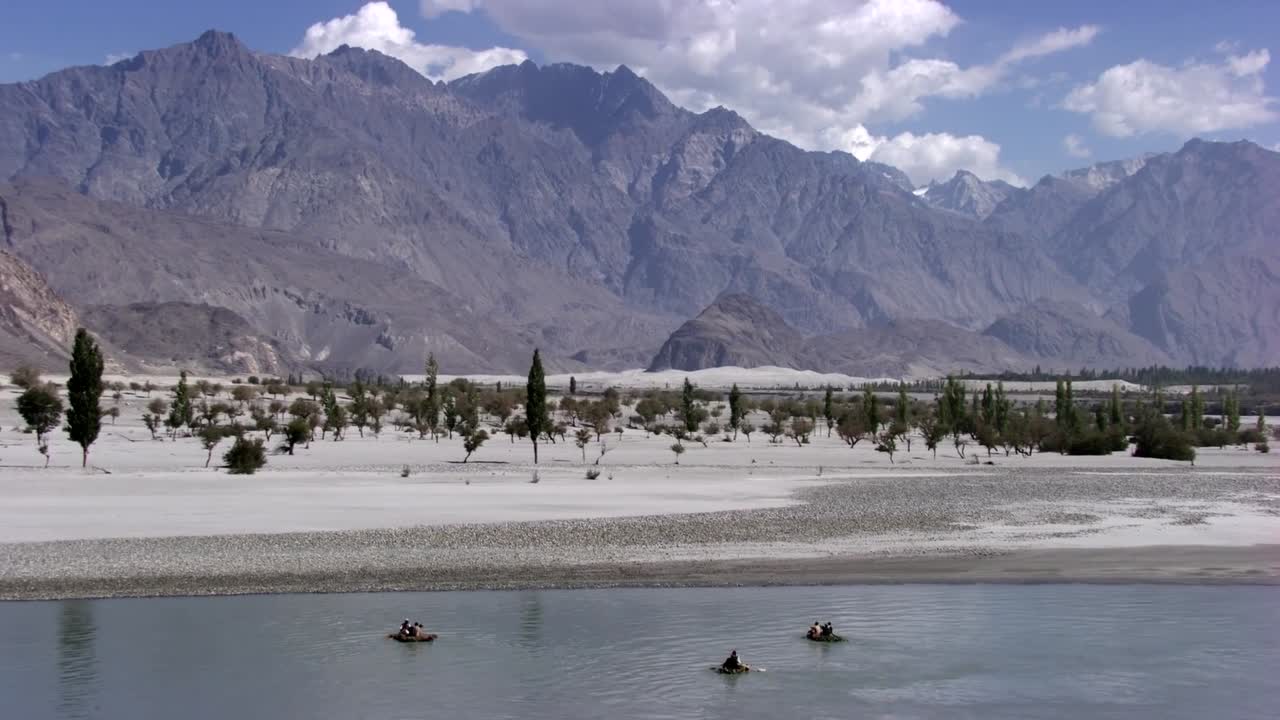 A serene aerial view of three small boats drifting across a calm river in a remote valley, framed by sandy plains and towering, jagged mountains. The peaceful water activity