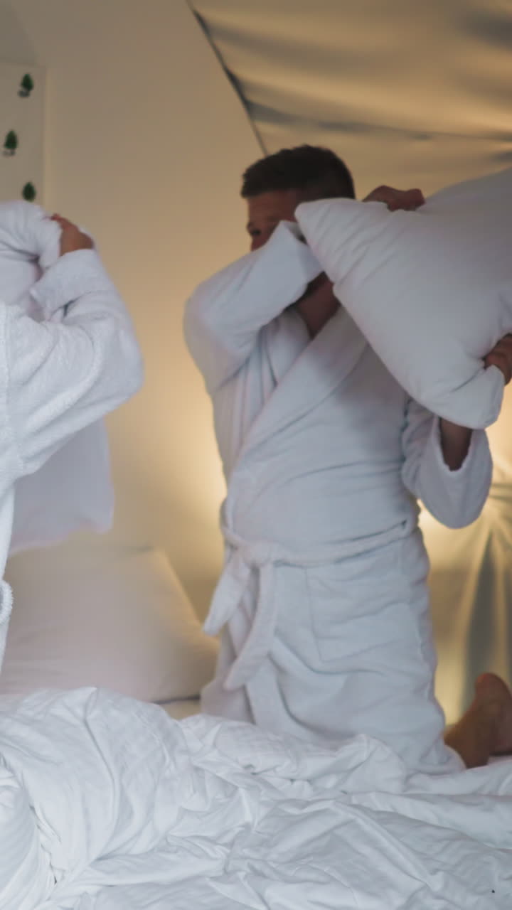 Woman falls on bed at pillow battle with boyfriend. Joyful young lovers enjoy playful fight and hug in glamping house. Newlywed passionate couple