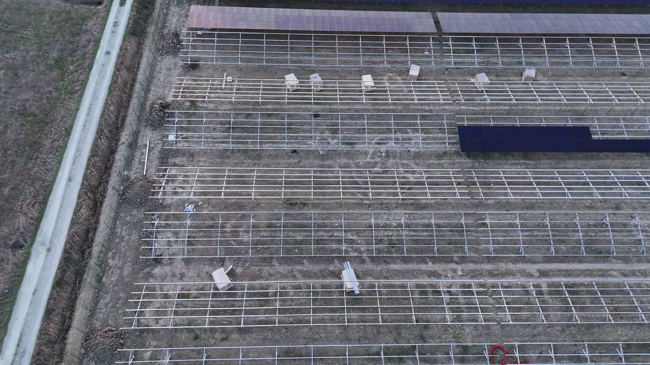 Struktur rangka fotovoltaik yang dipasang di tanah sedang dipasang dalam barisan teratur di seluruh lokasi konstruksi pembangkit listrik tenaga surya, dengan susunan yang sebagian sudah selesai terlihat, penampakan dari udara dalam gerakan lambat