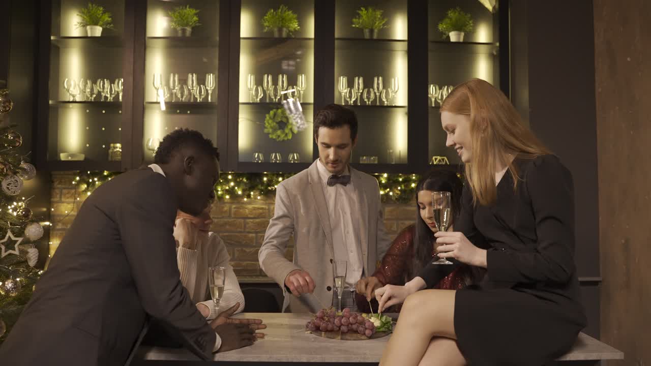 Group of friends dressed in elegant clothes celebrating the New year's party, they stand around the bar counter while drinking champagne and eating grapes