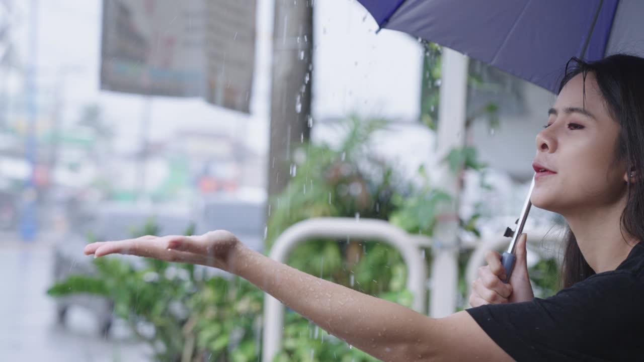 young asian female holding umbrella sit down waiting on the street side with hard pouring rain, rainy stormy season, reach her hand out touching rain drops, stuck in the rain, positive curios face