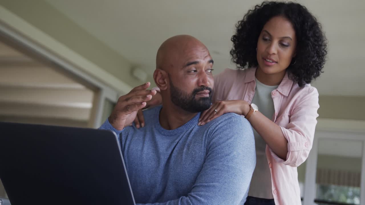Happy biracial couple doing paperwork and using laptop at table, slow motion