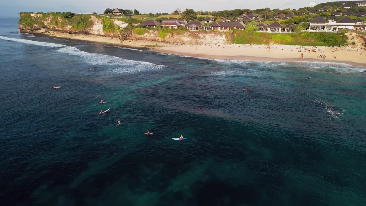 Cinematic drone view capturing surfers waiting for waves near Bali’s limestone cliffs, surrounded by crystal blue ocean and bright afternoon sunlight along the island’s peaceful southern coast