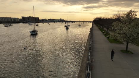 an aerial shot over sheepshead bay during a golden sunrise with boats anchored and a jogger running on the paved walkway along the water