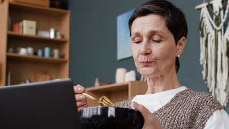 mature woman eating and looking at laptop at home