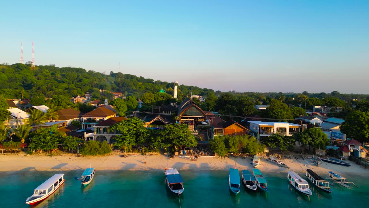 Drone video capturing a serene coastal town near Amed, Bali, featuring anchored boats and hilltop towers under clear blue skies. The panoramic view showcases vibrant colors and tranquil scenery