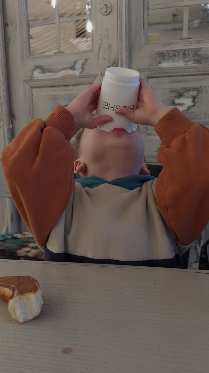 Caucasian toddler drinking from paper cup. Funny kid with foam moustache after drinking cocoa. Vertical video.