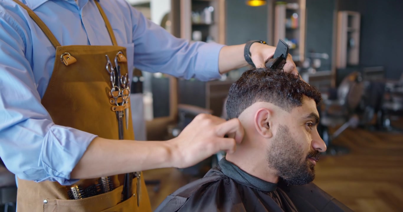 Mandlig frisør trimmer mandlig afrikansk kunde til en fade-klipning med klippemaskine, kam og forklæde i butikken, med plads til tekst.