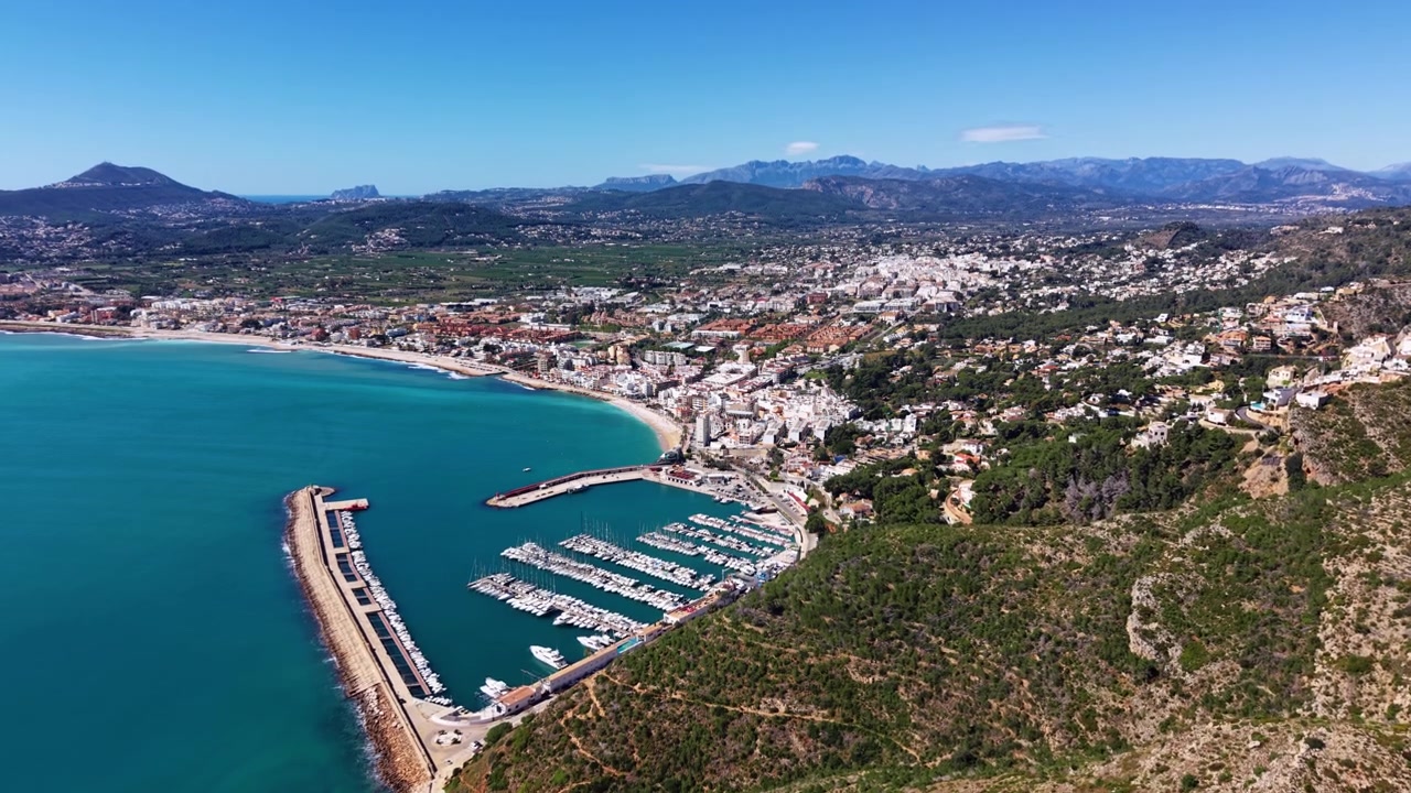 Città costiera di Javea sulla Costa Blanca spagnola con acque mediterranee turchesi, un porto turistico pieno di barche a vela, una spiaggia sabbiosa, edifici sulla collina e uno sfondo montuoso.