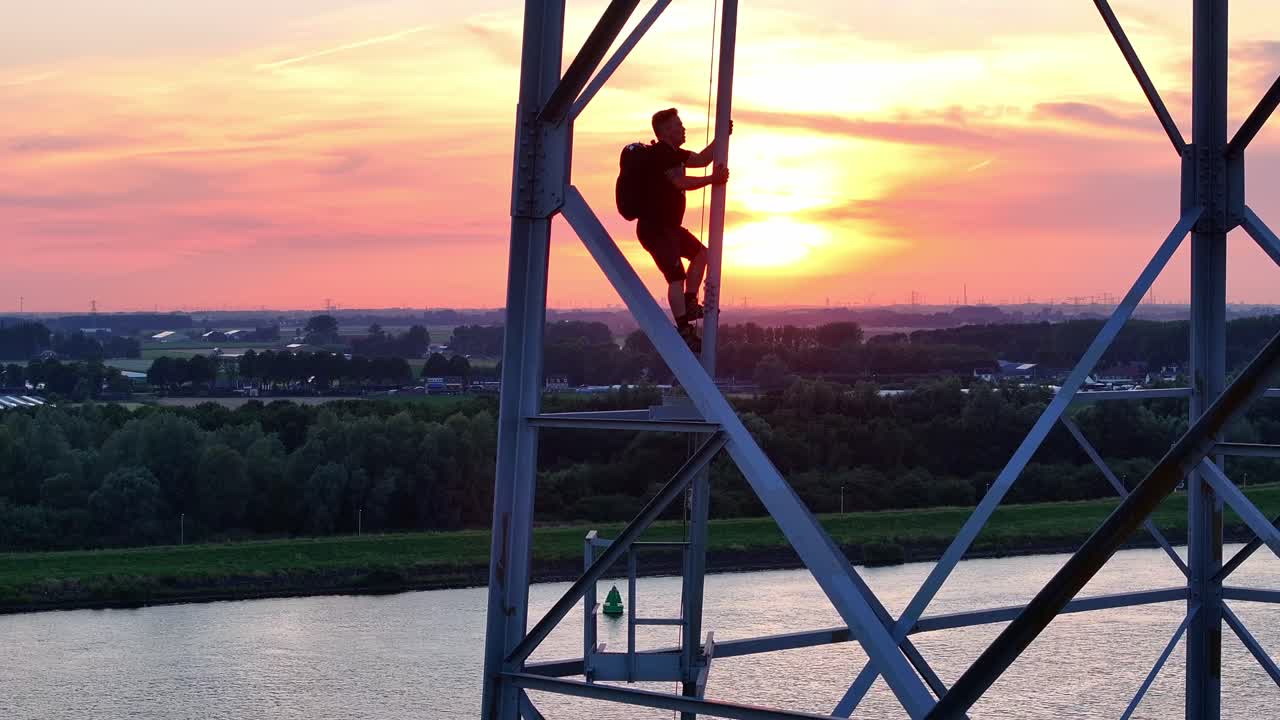 Person climbs a tall steel structure at sunset with a river and forest in the background