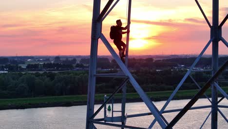 person climbs a tall steel structure at sunset with a river and forest in the background