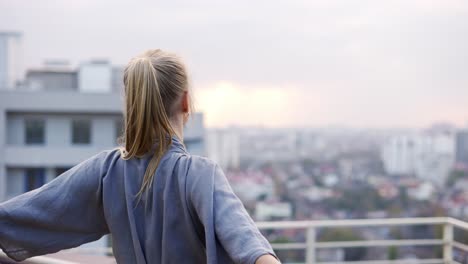 Mujer-Rubia-Disfruta-De-La-Vista-De-La-Ciudad-Y-Dobla-Su-Mano-En-Gesto-Namaste,-Vista-Trasera