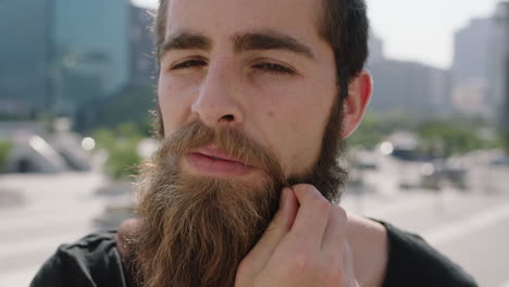 slow motion portrait of cute young confident hipster man student looking serious pensive at camera stroking beard in sunny urban city background