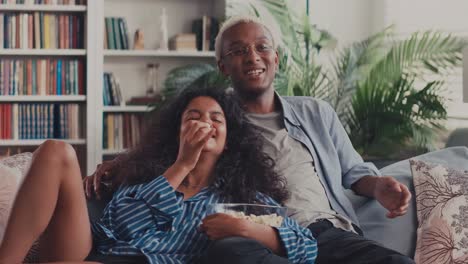 diverse couple laughing watching tv and eating popcorn sitting on sofa at home