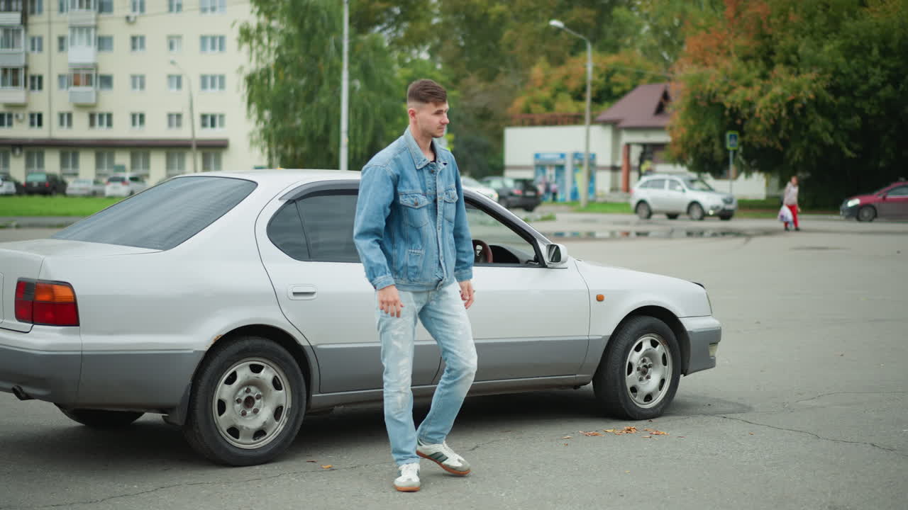 Un giovane esce da una berlina color argento a metà strada, controlla il telefono, indossa giacca e jeans di denim, apre la portiera e ispeziona il cruscotto, gesto di frustrazione e pausa, parcheggio di periferia con edifici e alberi, spontaneo