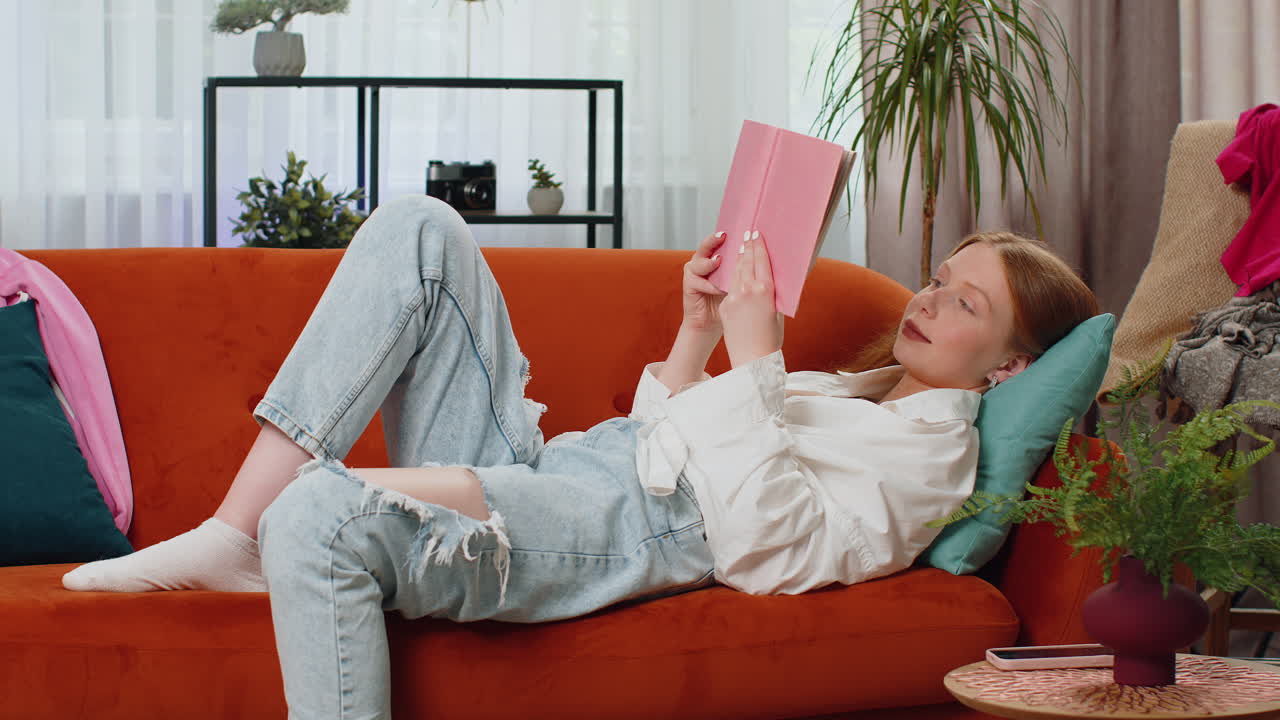 Teen girl reading interesting book turning pages smiling enjoying literature taking a rest on sofa
