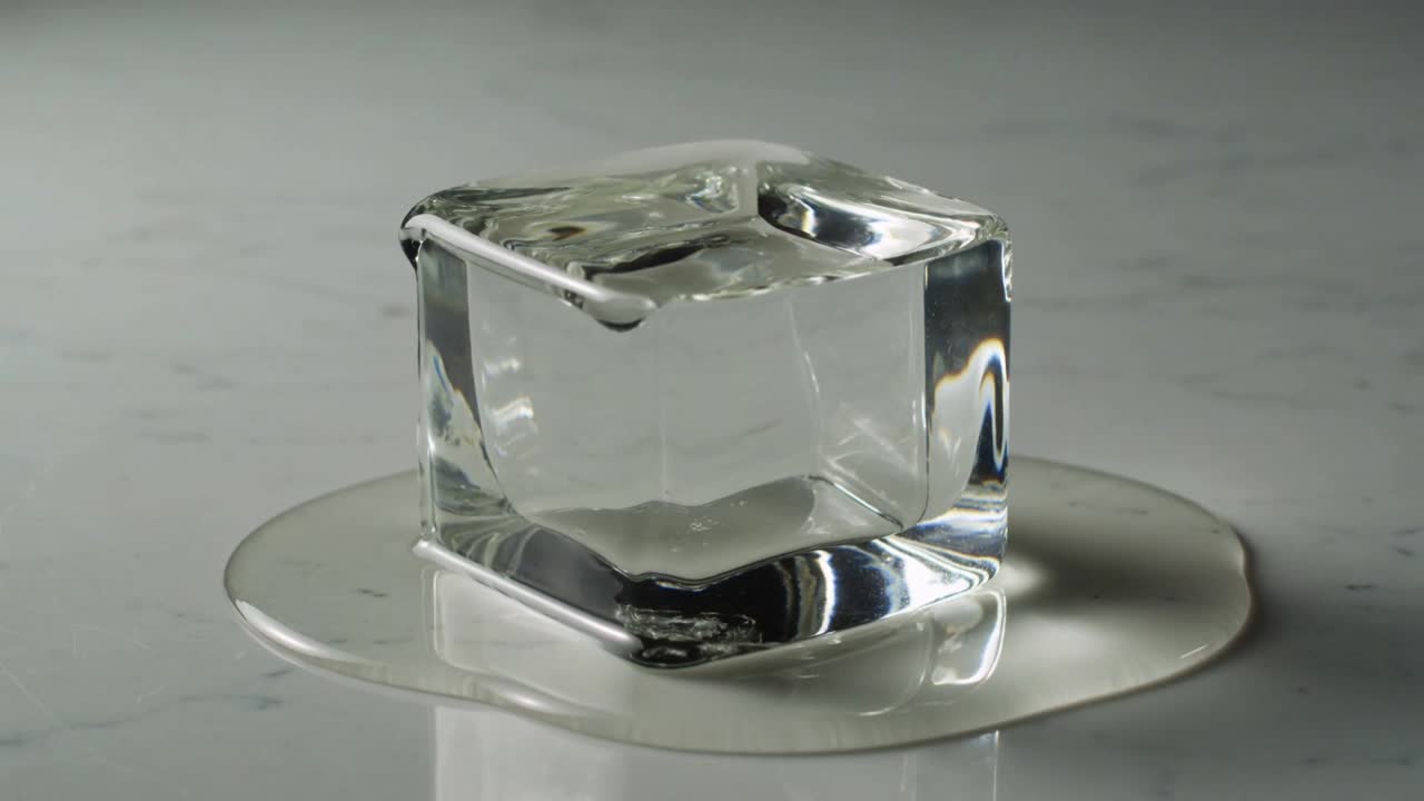 Un singolo cubetto di ghiaccio trasparente che si scioglie su un ripiano lucido, riscaldandosi e formando una pozzanghera, una gocciolina e un ruscello.