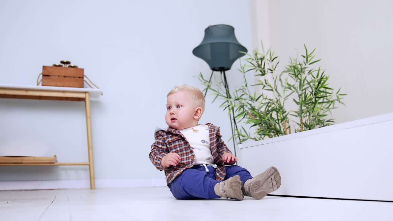 Lovely blond baby boy in checkered shirt sitting on the floor indoors. Cute one year old toddler starts to cry. Low angle view.